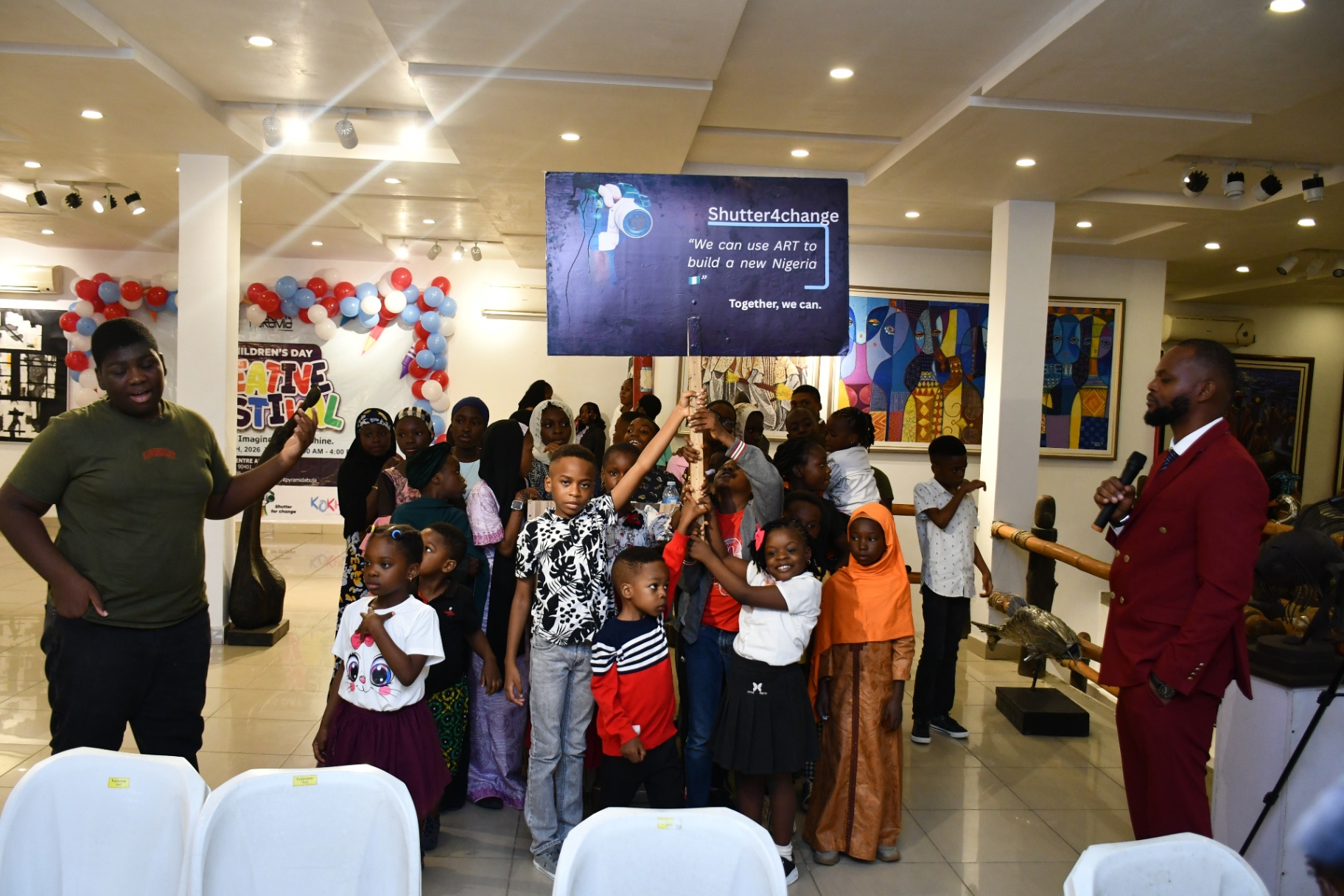 A wide view of children gathered under the Shutter4change placard with a host on either side